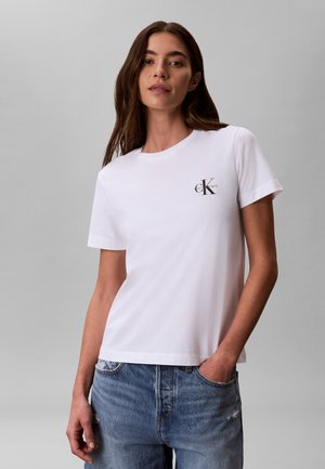 Žena nosiaca biele tričko Calvin Klein a modré džínsy, stojaca s jednou rukou v vrecku na jednoduchom sivom pozadí.