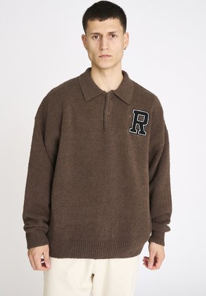 Ung mand iført en brun tekstureret langærmet pullover med krave og et sort-hvidt bogstav "R" patch på brystet.