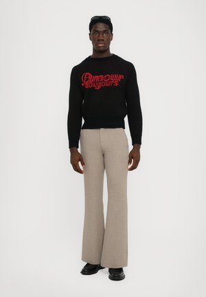 Black knitted sweater with red text, paired with beige flared trousers and black shoes. Model stands against a plain white background.