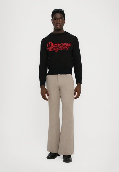 Maglione nero a maglia con testo rosso, abbinato a pantaloni svasati beige e scarpe nere. Modello in piedi contro uno sfondo bianco semplice.