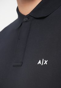 Close-up de uma pessoa a usar uma camisa polo negra com colarinho abotoado e um logo branco bordado "A|X" no peito.