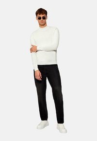 Weißer gestrickter Pullover mit langen Ärmeln und Rundhalsausschnitt, kombiniert mit schwarzen Jeans und weißen Sneakern. Das Model hat kurzes braunes Haar und eine Sonnenbrille.