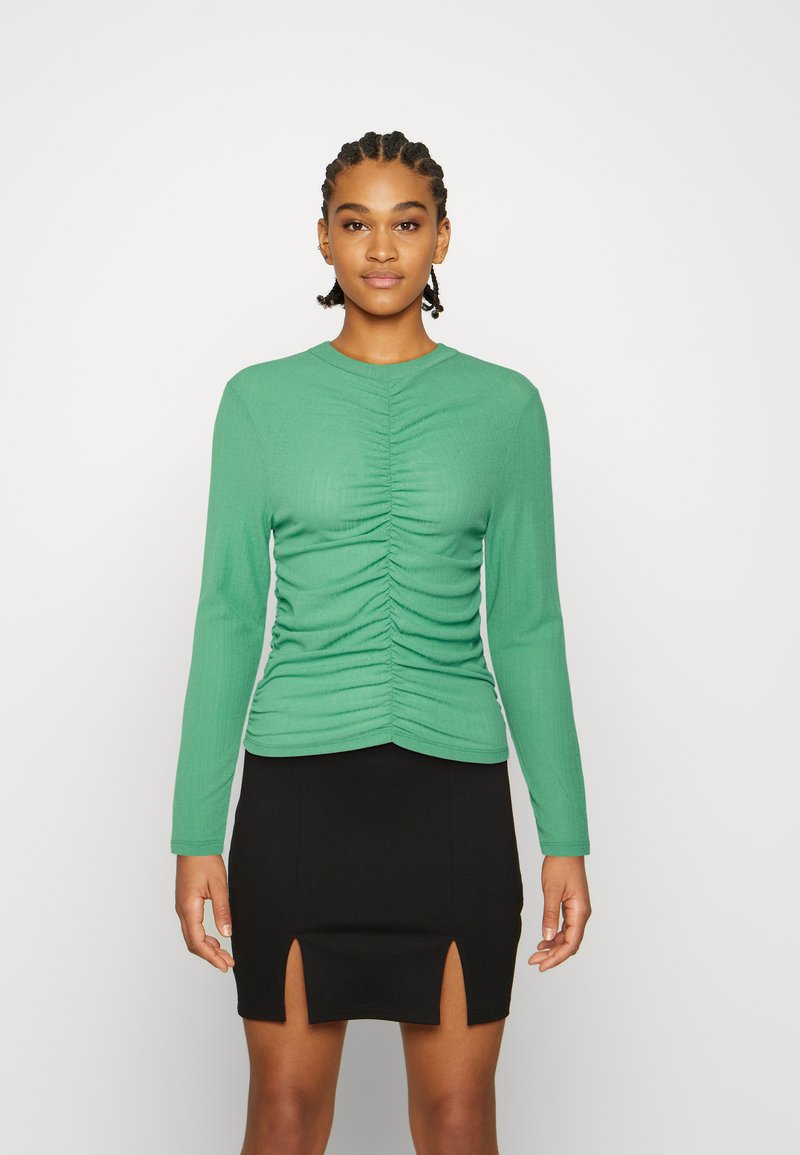 Top verde de manga longa com textura franzida na frente, combinado com uma saia preta com fendas frontais. Design ajustado e tecido canelado.