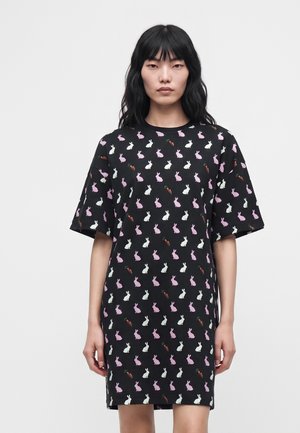 Femme portant une robe noire à manches courtes avec des motifs de lapins et de carottes roses et blancs, debout devant un fond clair uni.