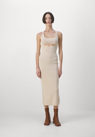 Beige fitted dress with a square neckline and cut-out detail below the bust. Made from a stretchy material, falling mid-calf. Brown heeled boots.