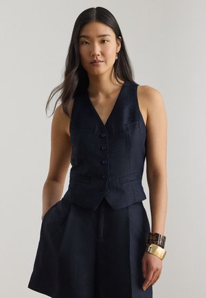 Γυναίκα με μακριά σκούρα μαλλιά, φορώντας αμάνικο μαύρο γιλέκο με κουμπιά και μαύρο σορτς που ταιριάζει, με δύο βραχιόλια στον δεξί της καρπό.