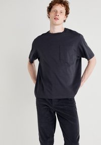 T-shirt en coton gris foncé à manches courtes, col rond et poche avant ; porté avec un pantalon sombre assorti, affichant une coupe décontractée.