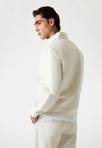 Cream jacket made of lightweight material, featuring a collar, full zipper, and ribbed cuffs. Paired with matching cream pants.