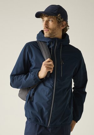 Uomo con giacca blu navy e cappello tiene la cinghia dello zaino grigio, guardando leggermente di lato su uno sfondo semplice.