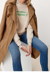 Trench-coat marron porté sur un sweat-shirt beige clair avec un texte vert "positive MINDSET", associé à un jean bleu. Les poignets du sweat-shirt présentent un motif.