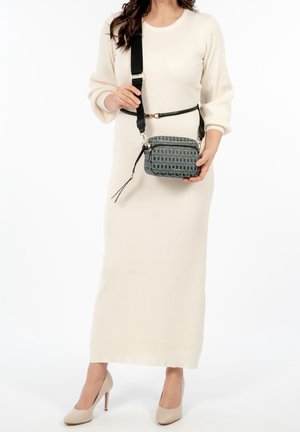 Femme vêtue d'une longue robe côtelée crème et de talons beiges, tenant un sac bandoulière bleu à motif avec une large sangle noire.