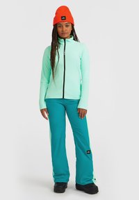 Mintgröna fleecejackan med svart dragkedja, ihop med tealbyxor med ljusgröna sidostripes. Orange stickad mössa på huvudet.