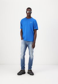 Camiseta azul de manga corta con un pequeño logo, combinada con jeans de un azul claro y zapatillas negras texturizadas. El modelo lleva gafas de sol reflectantes.