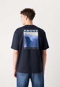 Tricou din bumbac bleumarin, cu mâneci scurte. Prezintă o grafică mare pe spate, cu textul "ABOVE" deasupra unei imagini abstracte a unui munte.
