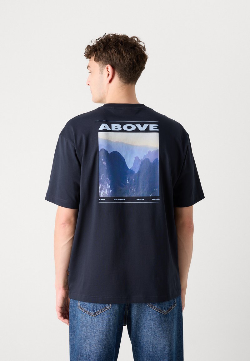 T-shirt in cotone blu navy con maniche corte. Presenta una grande grafica sul retro, con il testo "ABOVE" sopra un'immagine astratta di una montagna.