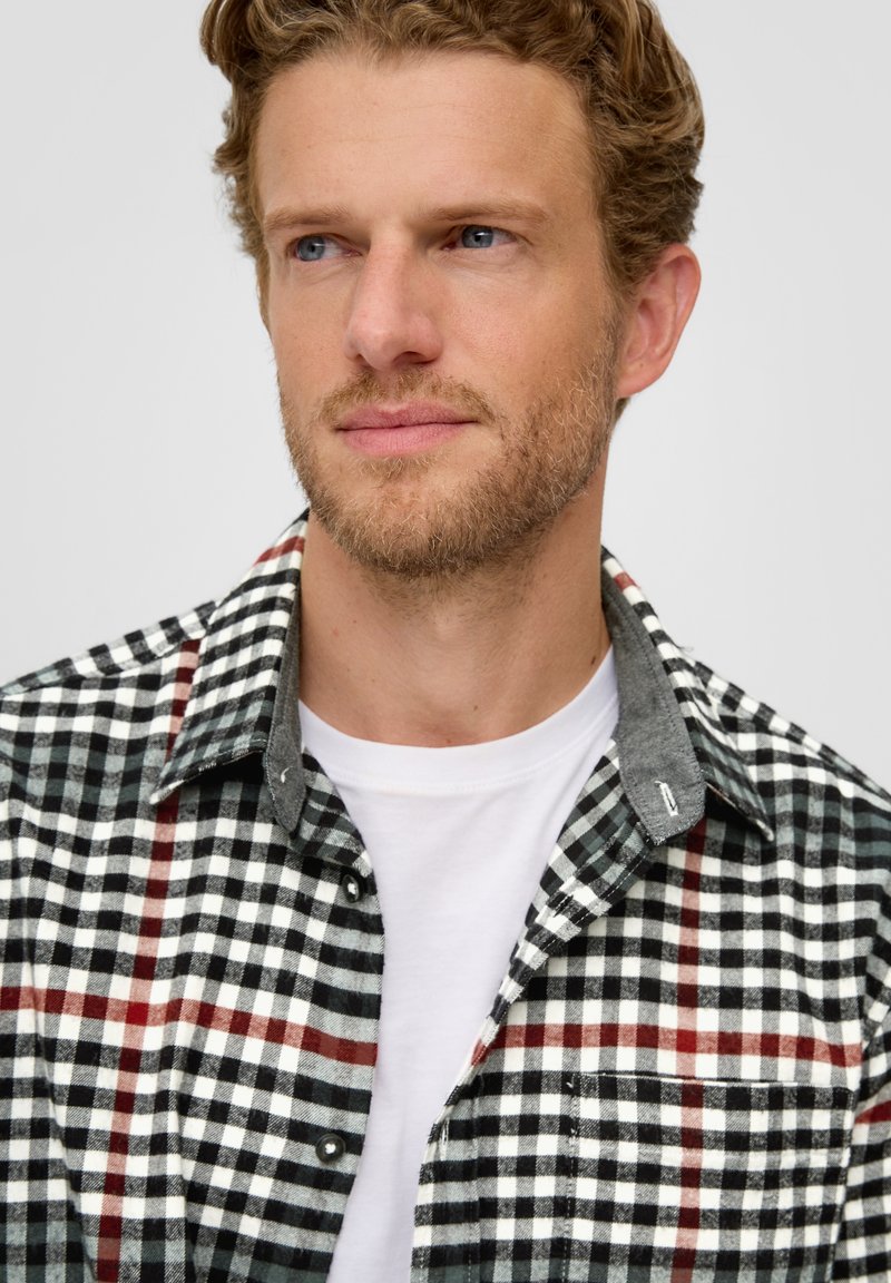 Czarna i biała koszula flanelowa w kraciasty wzór z czerwonymi akcentami, szarym kołnierzykiem i detalem kieszeni, noszona na białej koszulce.
