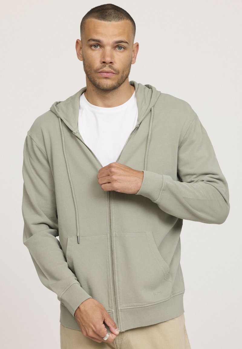 Sweat à capuche zippé vert clair avec une capuche à cordon de serrage, poches avant et poignets côtelés. Porté ouvert sur un t-shirt blanc. Texture de tissu douce.