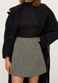 Zwarte turtleneck onder een lange zwarte jas, gecombineerd met een korte rok in een houndstooth-patroon in zwart en wit. Zachte stof, gestructureerde pasvorm.