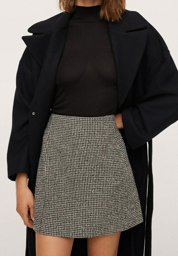 Zwarte turtleneck onder een lange zwarte jas, gecombineerd met een korte rok in een houndstooth-patroon in zwart en wit. Zachte stof, gestructureerde pasvorm.
