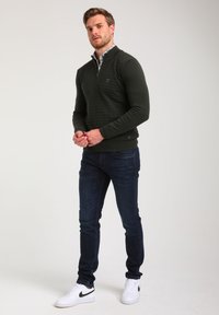 Tamsiai žalias megztinis su deimantiniu raštu, su užtrauktuku ant kaklo, dėvimas virš raštuoto marškinėlio. Suderintas su tamsiais džinsais ir baltais sportbačiais.