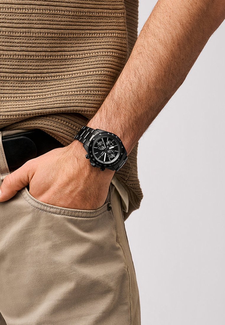 Reloj de muñeca negro con brazalete de acero inoxidable, esfera redonda, subesferas y acentos luminosos, usado por una persona con una parte superior texturizada de color marrón.