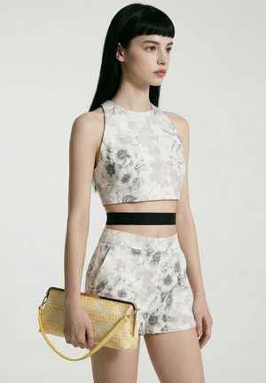 Jeune femme aux longs cheveux noirs portant un crop top blanc à motifs floraux et un short, tenant une pochette jaune texturée.