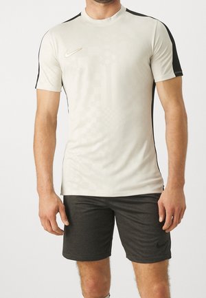 Homme portant un maillot de sport beige clair avec des bandes noires sur les épaules et un short de sport gris foncé sur un fond uni.