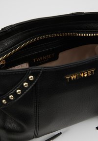 Borsa in pelle nera con hardware dorato, dotata di chiusura a zip, tasca interna e dettagli in borchie sui lati. Etichetta del marchio visibile all'interno.