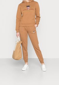 Felpa marrone con logo, pantaloni jogger abbinati e scarpe da ginnastica bianche. Il modello tiene una borsa marrone chiaro. Tessuto morbido, vestibilità casual, polsini a coste.