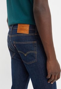 Mörkblå denimjeans med gula sömmar, med en läderlapp märkt "Levi Strauss & Co" ovanför bakfickan, slim fit-design.