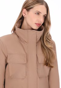 Beige jakke med høj krave, to frontlommer med klapper og en glat tekstur. Designet med en lynlåslukning foran.