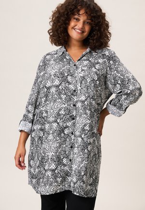 Glimlachende vrouw met krullend haar, gekleed in een zwart-witte, lange mouwen tuniek met een bloemenkantpatroon en zwarte broek.