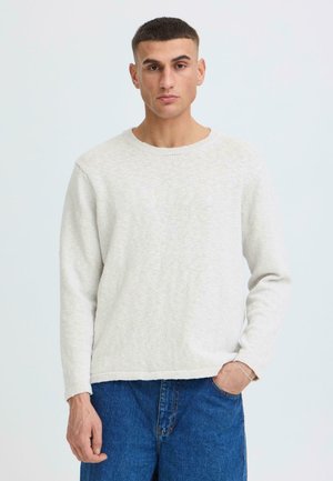 Jeune homme debout portant un pull en maille beige clair et un jean bleu, une main dans la poche, sur un fond uni.