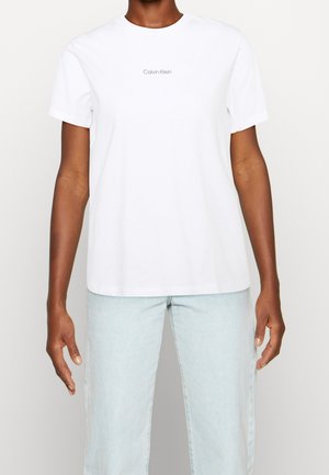 Persoon draagt een eenvoudige witte Calvin Klein T-shirt met korte mouwen en lichtblauwe spijkerbroek, staand tegen een witte achtergrond.
