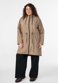 Beiger langer Mantel mit verstellbarer Taille, vorderem Reißverschluss und Kapuze. Schwarze Kordeln und Taschen sorgen für Funktionalität. Kombiniert mit schwarzen weiten Hosen.