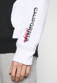 Sweat-shirt à capuche avec des blocs de couleur noir et blanc, arborant un texte en damier "CALIFORNIA" et des accents rouges sur la manche. Tissu doux.