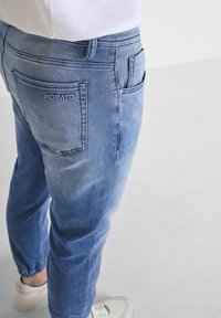 Pantalones jogger de denim azul claro con puños elásticos, que cuentan con un bolsillo trasero visible con el logotipo bordado "MORATO". Textura suave, corte relajado.