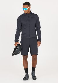 Schwarze Radsportjacke und -shorts aus dehnbarem Stoff. Die Jacke hat einen Reißverschluss; die Shorts haben Taschen. Schwarze Radsportschuhe und ein Helm sind ebenfalls enthalten.