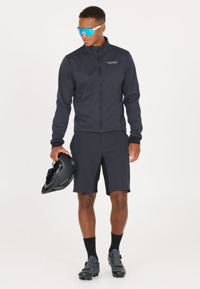 Schwarze Radsportjacke und -shorts aus dehnbarem Stoff. Die Jacke hat einen Reißverschluss; die Shorts haben Taschen. Schwarze Radsportschuhe und ein Helm sind ebenfalls enthalten.