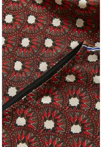 Motifs géométriques en forme d'étoile rouges, noirs et beiges sur tissu avec une fermeture éclair diagonale noire et une petite languette en tissu rayé.