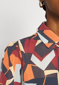 Kurzärmlige Bluse mit einem geometrischen Muster in Orange, Rot, Marineblau und Beige. Glatte Textur mit Kragen und Knopfdetails.