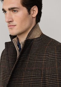 Abrigo de lana de cuadros marrón con un alto cuello, que presenta un cuello beige en contraste y un cierre de cremallera en la parte delantera. Tejido texturizado con patrones visibles.