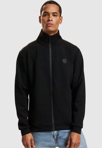 Sort zip-jakke lavet af fleece, med høj krave, lynlåset front og et hexagonalt logo på brystet. Elastiske manchetter og kant.