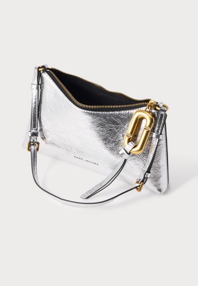Petit sac à main argenté Marc Jacobs avec fermeture éclair dorée et détail de fermoir doré, partiellement ouvert avec une fine bandoulière sur fond blanc.