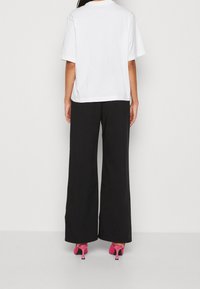 Kvinna som bär en lös vit kortärmad skjorta, vida svarta byxor och rosa högklackade sandaler, stående med ryggen mot kameran.