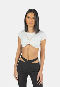 Top bianco corto con design frontale attorcigliato, abbinato a pantaloni neri a vita alta con dettagli a taglio e una collana in oro.