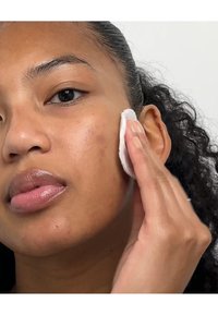 Persoon gebruikt een wattenpad om het gezicht te reinigen. De huid oogt glad met lichte oneffenheden. Het haar is krullend en naar achteren gekamd.