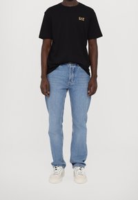 Camiseta de algodón negra con un pequeño logo dorado, combinada con unos vaqueros de mezclilla azul claro y zapatillas blancas, con cierre de cordones y textura suave.