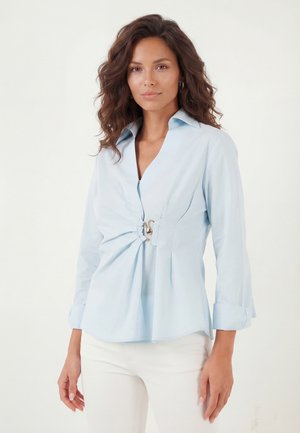 Kvinde med bølget brunt hår iført en lyseblå bluse med lange ærmer og en gylden lås i midten samt hvide bukser, står foran en ensartet baggrund.