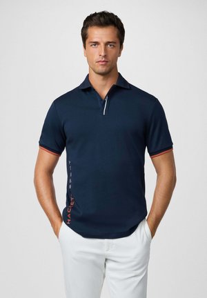 Marineblaues Poloshirt mit einem Kragen mit zwei Knöpfen, weißen Reißverschlusdetails und roten Akzenten an den Bündchen und an der Seite. Mit Logodruck an der Seite.